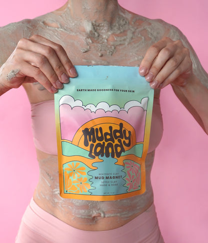 Woman covered in Muddyland Bentonite Clay Bath Mud Mask holding Mud Magnet packaging, showcasing the detox clay soak against a pink background.