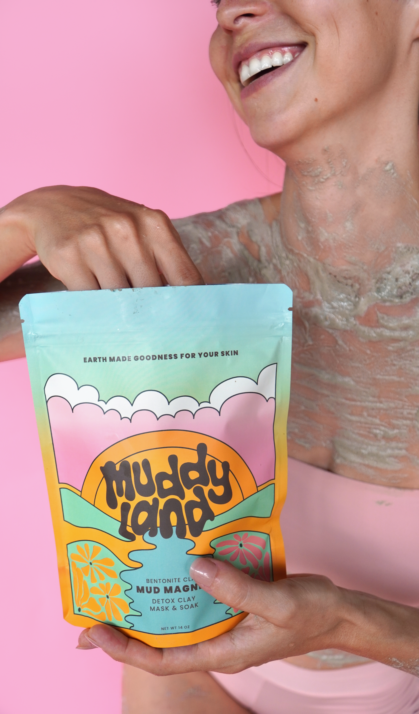 Woman smiling and applying Muddyland Mud Magnet detox bath with bentonite clay in a mud mask while holding the product package against a pink background.